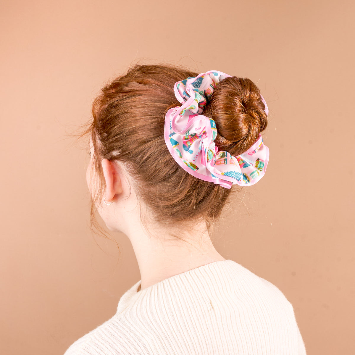A Sugarplum Lane Ruffle Up Scrunchie in pink with a festive pattern of nutcrackers and Christmas trees worn in a hair bun.