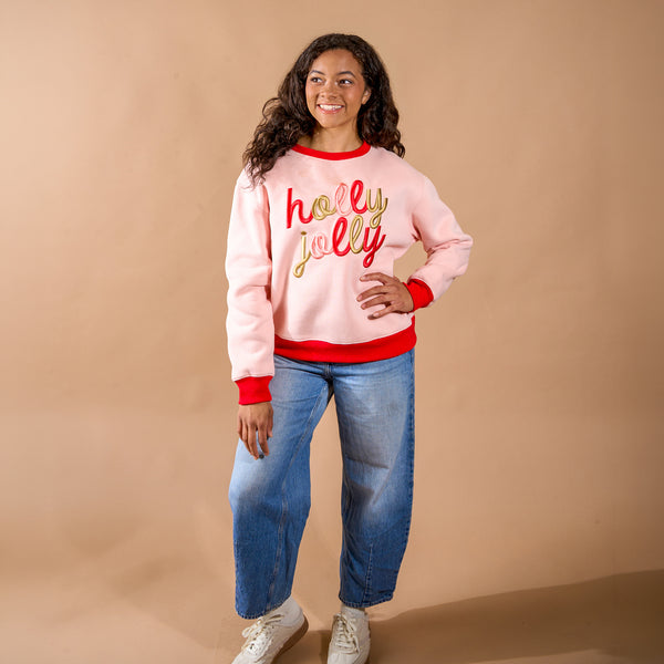 A smiling person wears a pale pink Holly Jolly Signature Crew with red and gold lettering, red trim at the neck, cuffs, and hem, and blue jeans.