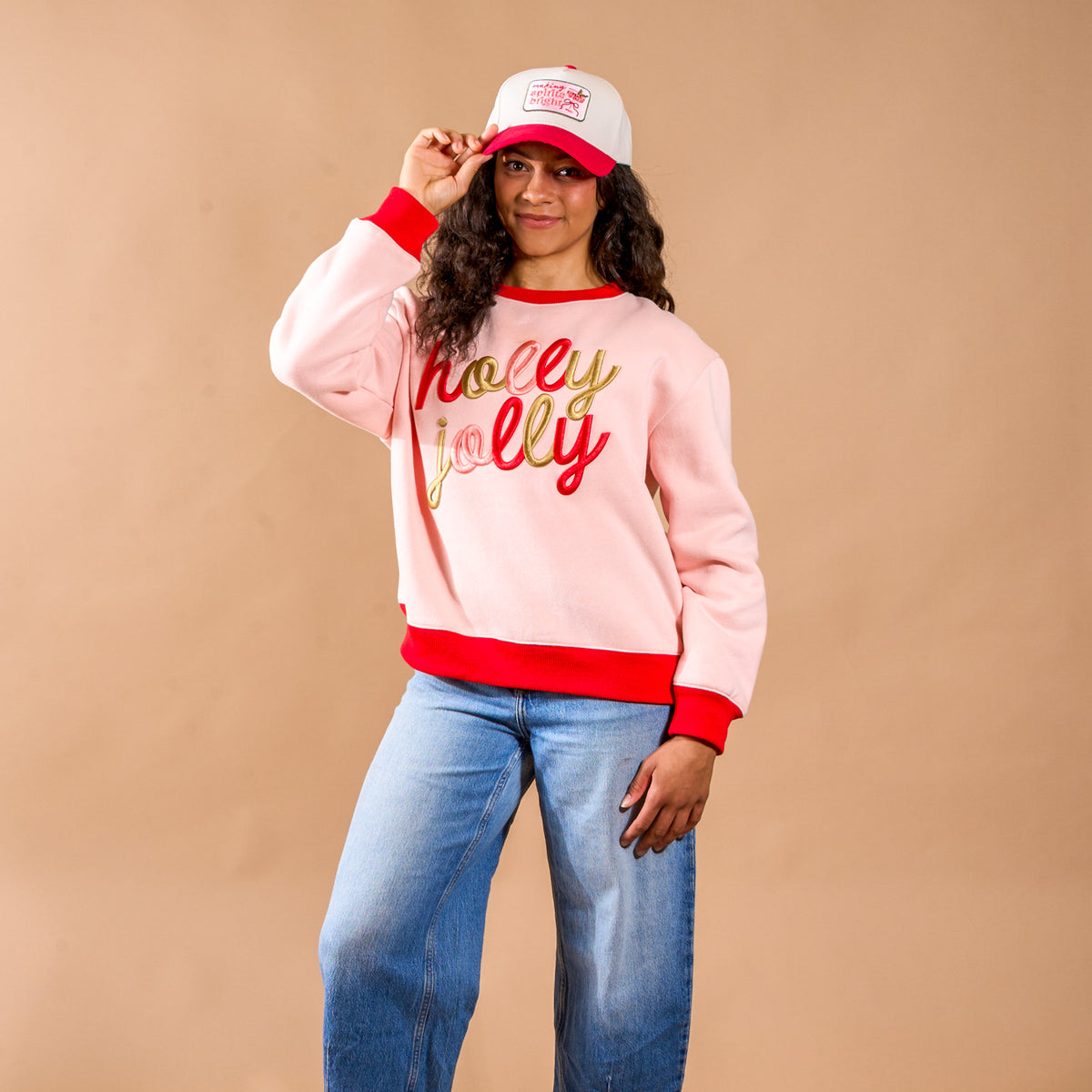 A person wears a light pink Holly Jolly Signature Crew sweatshirt with red and gold script across the chest, paired with blue jeans and a white and red baseball cap.