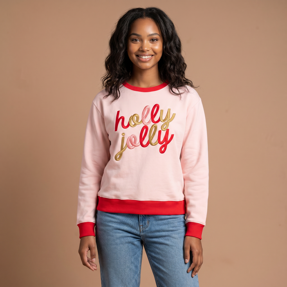 A person wears a light pink Holly Jolly Signature Crew sweatshirt with red trim and holly jolly embroidered in red and gold script on the chest.