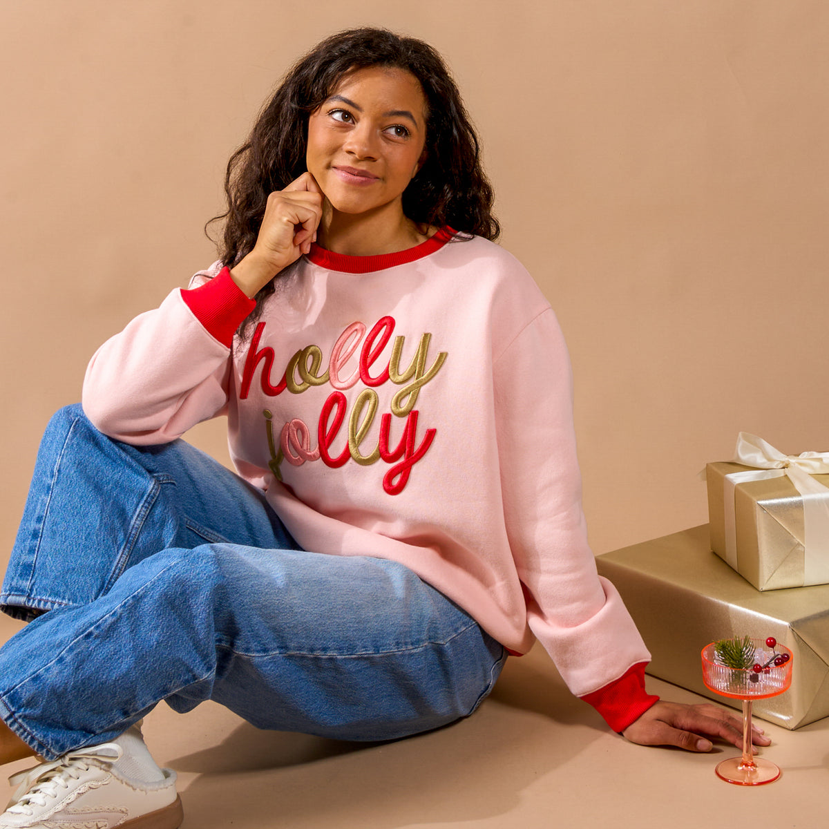 A person in a light pink Holly Jolly Signature Crew sweatshirt with holly jolly in red and gold script, sitting next to gift-wrapped boxes and a stemmed glass.