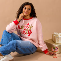 A person in a light pink Holly Jolly Signature Crew sweatshirt with holly jolly in red and gold script, sitting next to gift-wrapped boxes and a stemmed glass.