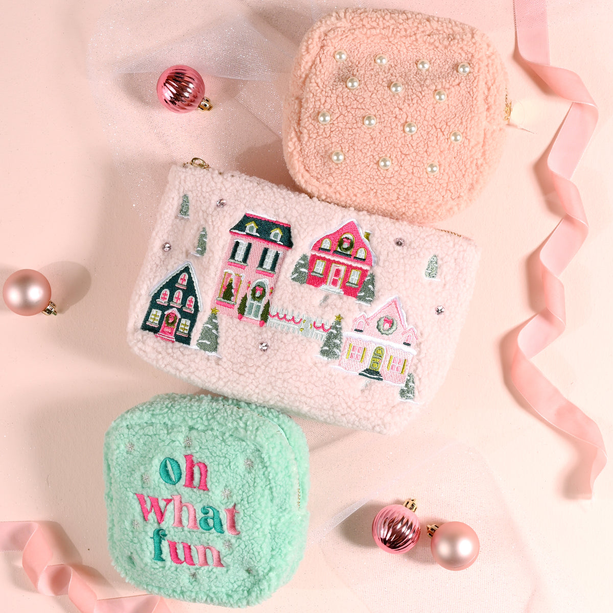 Three fuzzy pouches from The Darling Effect: a pink square Pink Pine Village Teddy Pouch with pearls, a larger rectangular one with an embroidered winter village scene, and a small mint green square one with the text oh what fun.