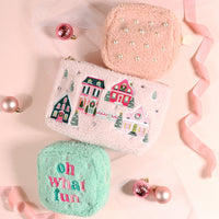 Three fuzzy pouches from The Darling Effect: a pink square Pink Pine Village Teddy Pouch with pearls, a larger rectangular one with an embroidered winter village scene, and a small mint green square one with the text oh what fun.