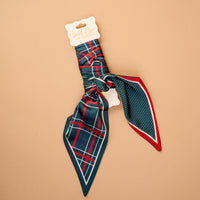 The Evergreen Plaid Twirl & Tie Scarf Accessory by The Darling Effect is a green, red, and white plaid satin scarf tied in a knot, with one tail showing a small white polka dot pattern on a green background.