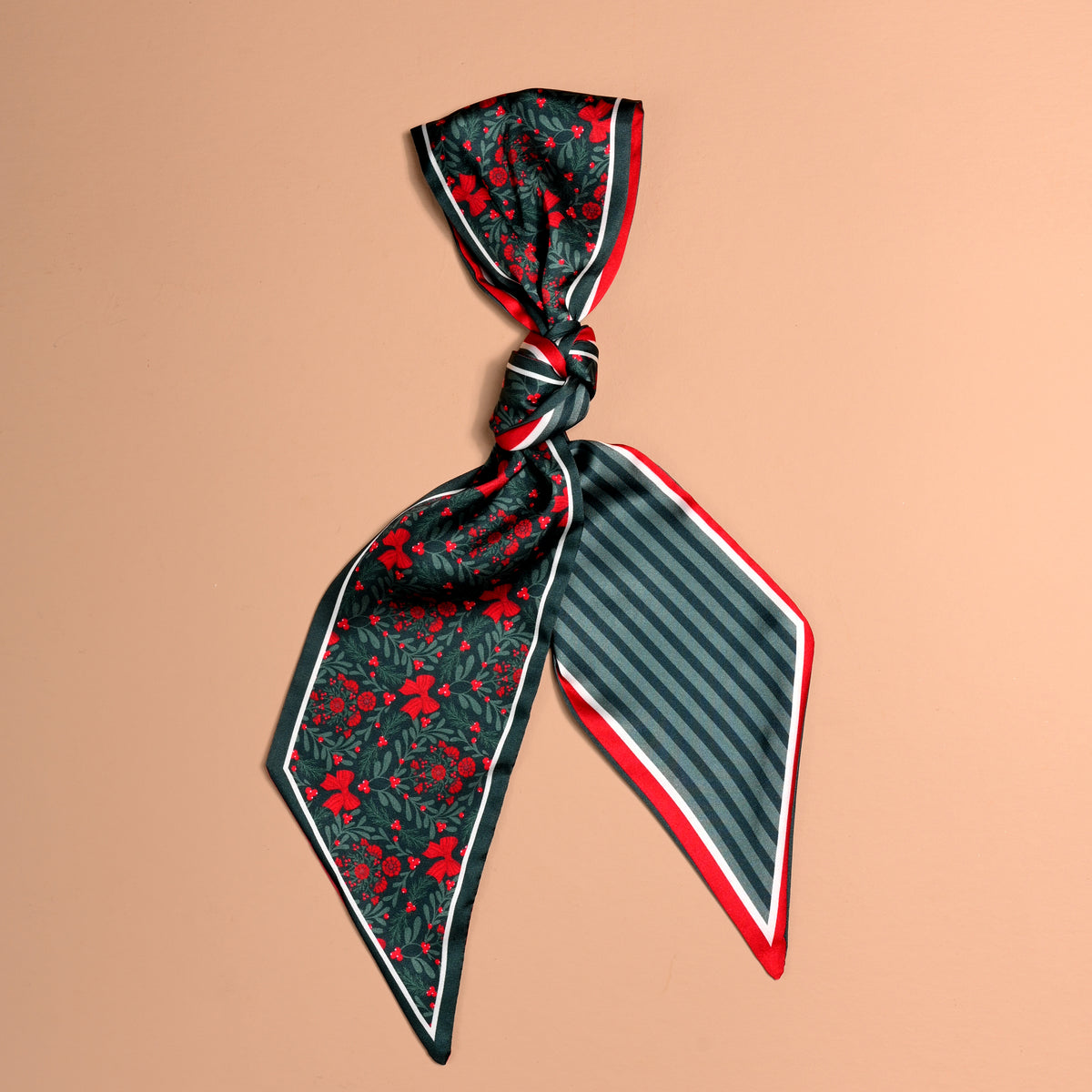 A Merry Moment Twirl & Tie Scarf Accessory, a long, narrow satin scarf tied in a knot, featuring a dark green floral print on one side and dark green stripes on the other, both finished with a red and white border.