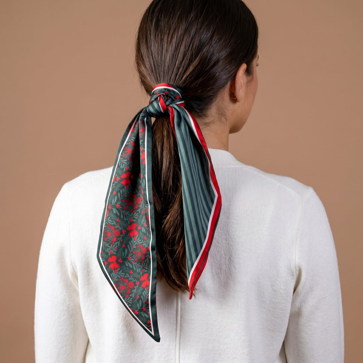 A Merry Moment Twirl & Tie Scarf Accessory tied in a ponytail, featuring a festive red and green floral print on one side and green stripes on the other for versatile styling.