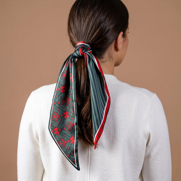A Merry Moment Twirl & Tie Scarf Accessory tied in a ponytail, featuring a festive red and green floral print on one side and green stripes on the other for versatile styling.