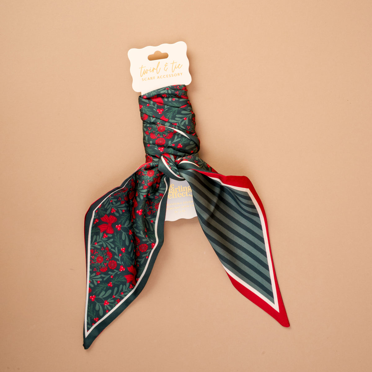 A Merry Moment Twirl & Tie Scarf Accessory by The Darling Effect, featuring a satin scarf with a green holiday holly and flower pattern on one side and dark green stripes with a red border on the other, displayed on a branded hanging card.