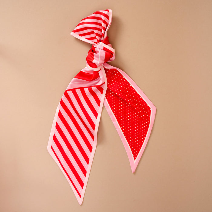 The Peppermint Stripes Twirl & Tie Scarf Accessory by The Darling Effect is a red and pink satin scarf tied in a knot, featuring one side with diagonal stripes and the other with a white polka dot pattern.