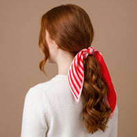 A ponytail tied with a Peppermint Stripes Twirl & Tie Scarf Accessory by The Darling Effect, featuring red and pink satin with striped and polka dot patterns.