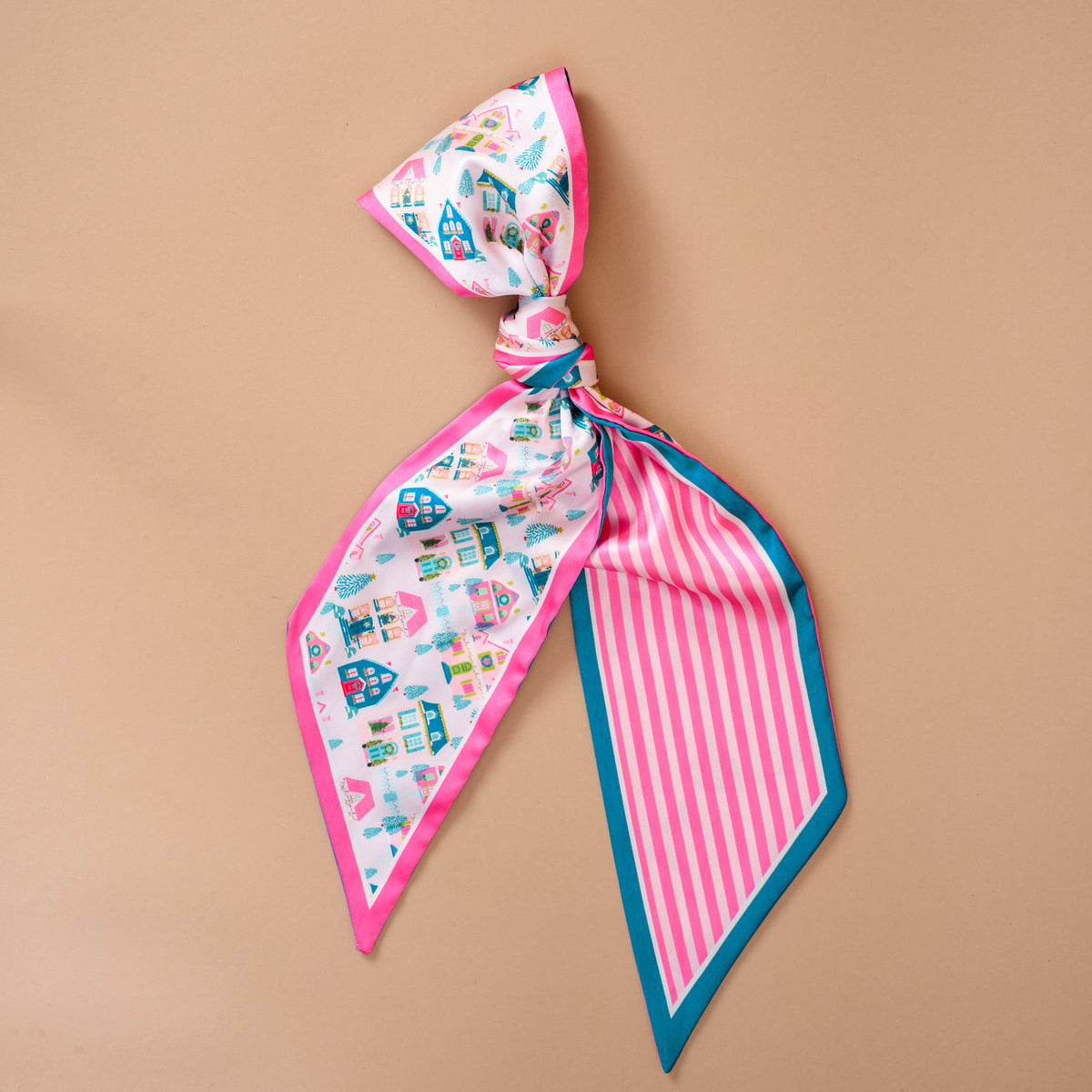 The Darling Effect Sugarplum Lane Twirl & Tie Scarf Accessory is a tied satin scarf with a pattern of colorful houses and trees on one side, pink and white stripes on the other, and teal trim.