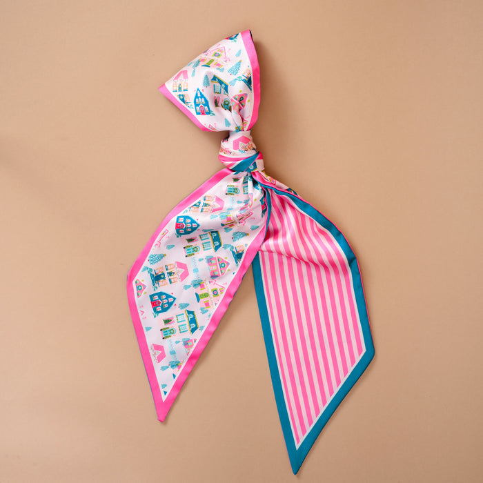 The Darling Effect Sugarplum Lane Twirl & Tie Scarf Accessory is a tied satin scarf with a pattern of colorful houses and trees on one side, pink and white stripes on the other, and teal trim.