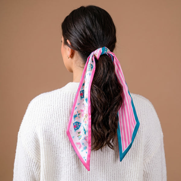 A person wearing a Sugarplum Lane Twirl & Tie Scarf Accessory tied around a ponytail, featuring a print of houses on one side and pink and white stripes on the other for versatile styling options.