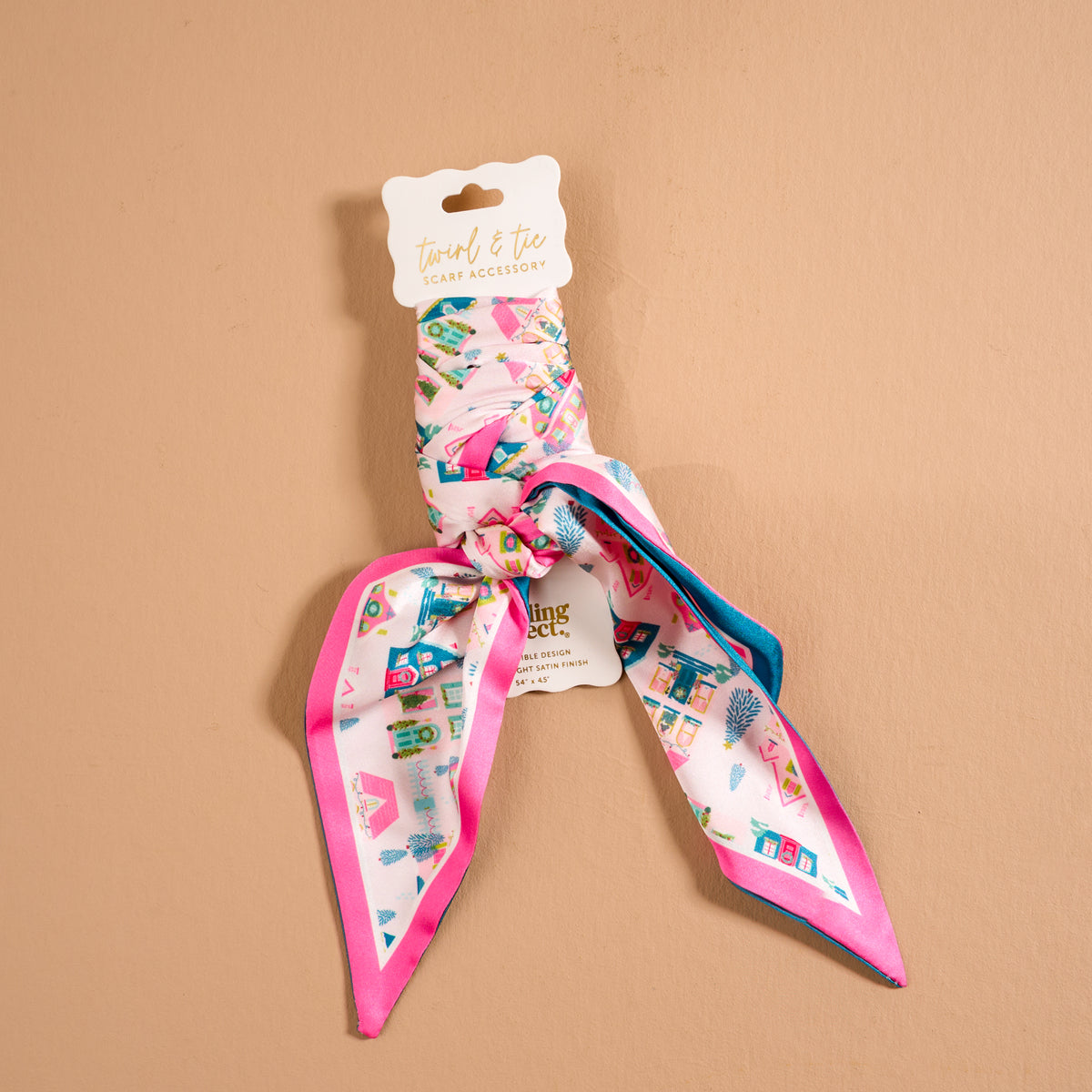 The Darling Effect Sugarplum Lane Twirl & Tie Scarf Accessory, a pink satin scarf with a whimsical pattern of small houses, trees, and stars, tied in a knot on a white product hang tag.