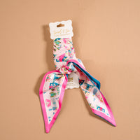The Darling Effect Sugarplum Lane Twirl & Tie Scarf Accessory, a pink satin scarf with a whimsical pattern of small houses, trees, and stars, tied in a knot on a white product hang tag.
