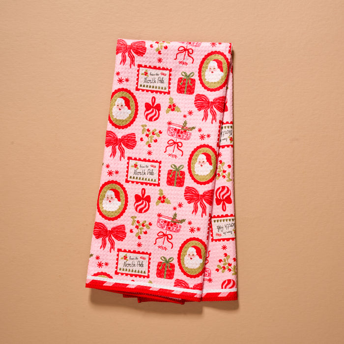 A folded pink Mistletoe Magic Waffle Tea Towel by The Darling Effect, featuring a repeating Christmas pattern of Santa Claus faces, gift boxes, red bows, and From the North Pole labels.