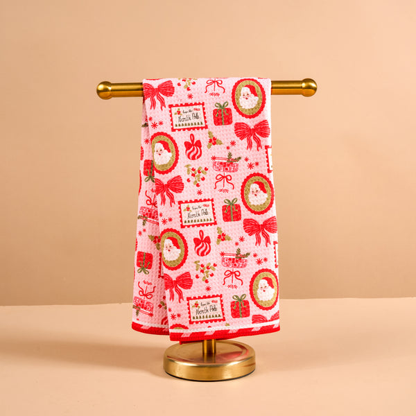 The Darling Effect Mistletoe Magic Waffle Tea Towel is a white cotton towel with a waffle texture, a red hem, and a repeating Christmas pattern of Santa faces, gifts, and mistletoe draped over a gold stand.