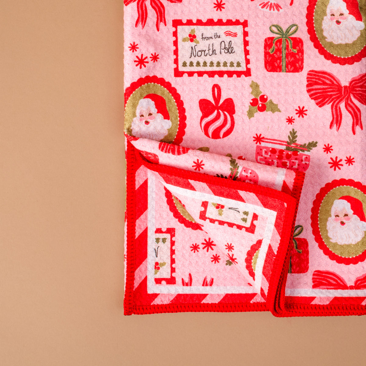 A Mistletoe Magic Waffle Tea Towel by The Darling Effect folded to show a pink pattern of Santa Claus faces, gifts, bows, and From the North Pole text, with a red-and-white candy cane striped border.