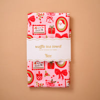 A pink Mistletoe Magic Waffle Tea Towel by The Darling Effect featuring a repeating holiday pattern of Santa Claus, gift boxes, and From the North Pole postage stamps, wrapped in a paper band.