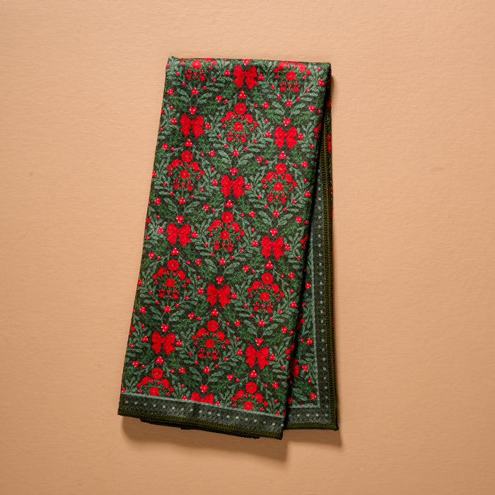 A Merry Moment Waffle Tea Towel, folded and featuring a festive green and red pattern of holly, berries, and ribbons on a solid tan background.