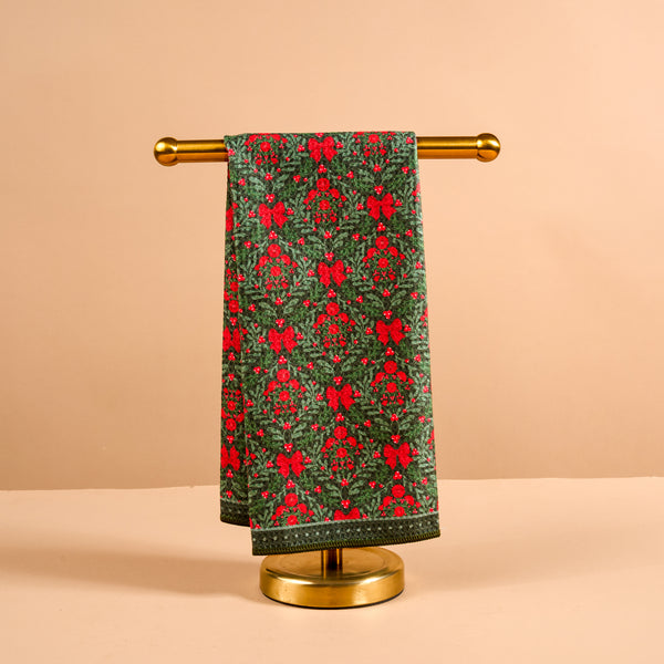 A Merry Moment Waffle Tea Towel in a waffle-weave cotton with a green botanical print featuring red bows and berries, hanging on a gold metal stand.