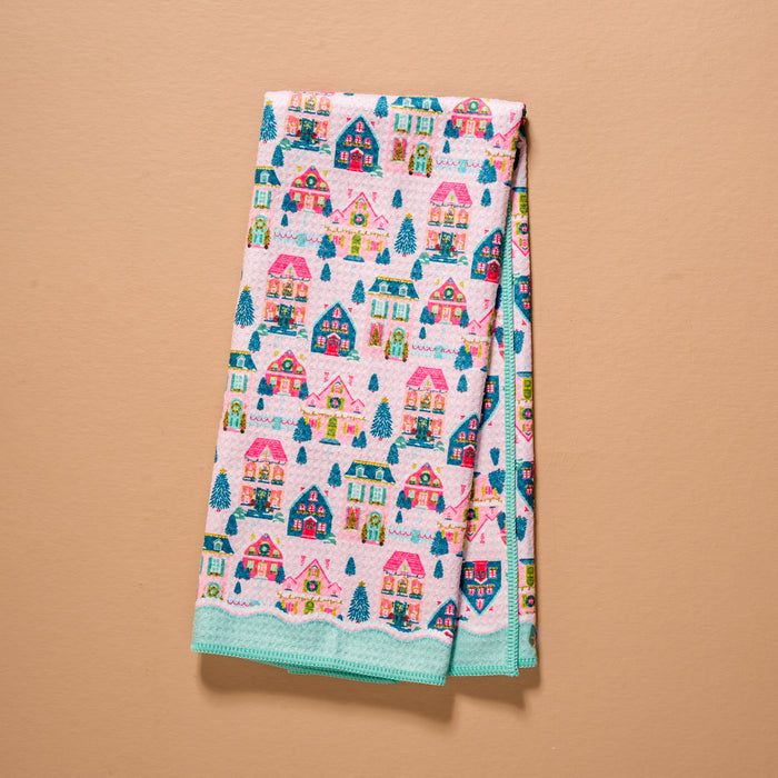 Sugarplum Lane Waffle Tea Towel folded to show a repeating pattern of colorful houses and evergreen trees on a pink background with a solid teal border.