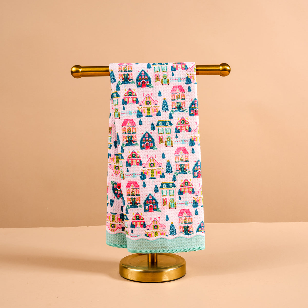 Sugarplum Lane Waffle Tea Towel featuring a repeating print of colorful holiday cottages and evergreen trees hanging on a gold towel stand.