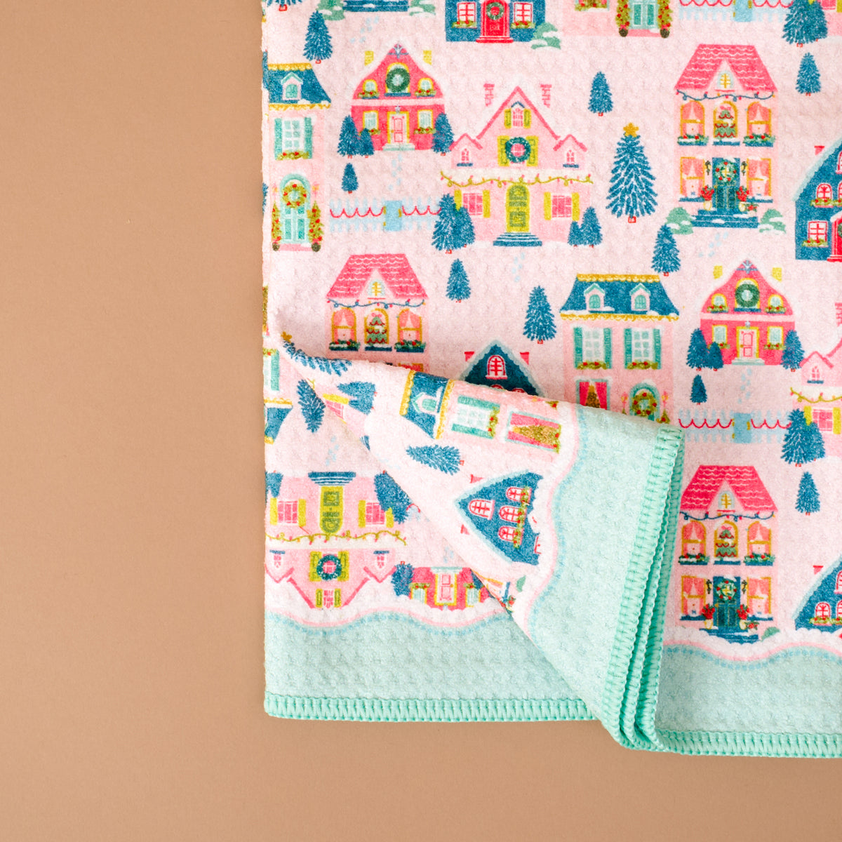 A folded Sugarplum Lane Waffle Tea Towel by The Darling Effect featuring a repeating pattern of colorful holiday houses, wreaths, and evergreen trees on a pink background with a solid mint-green border.