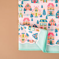 A folded Sugarplum Lane Waffle Tea Towel by The Darling Effect featuring a repeating pattern of colorful holiday houses, wreaths, and evergreen trees on a pink background with a solid mint-green border.
