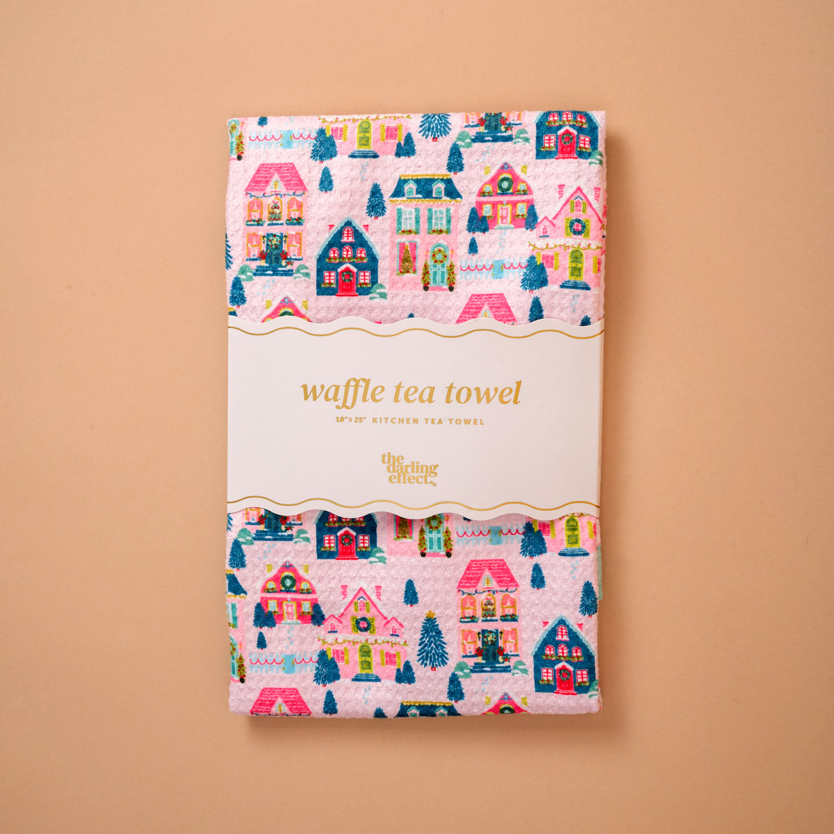 A folded pink Sugarplum Lane Waffle Tea Towel by The Darling Effect, featuring a colorful holiday house and pine tree pattern and wrapped in a paper band.