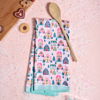 A folded Sugarplum Lane Waffle Tea Towel by The Darling Effect featuring colorful illustrated houses and trees, resting on a pink surface next to a wooden spoon and holiday cookies.