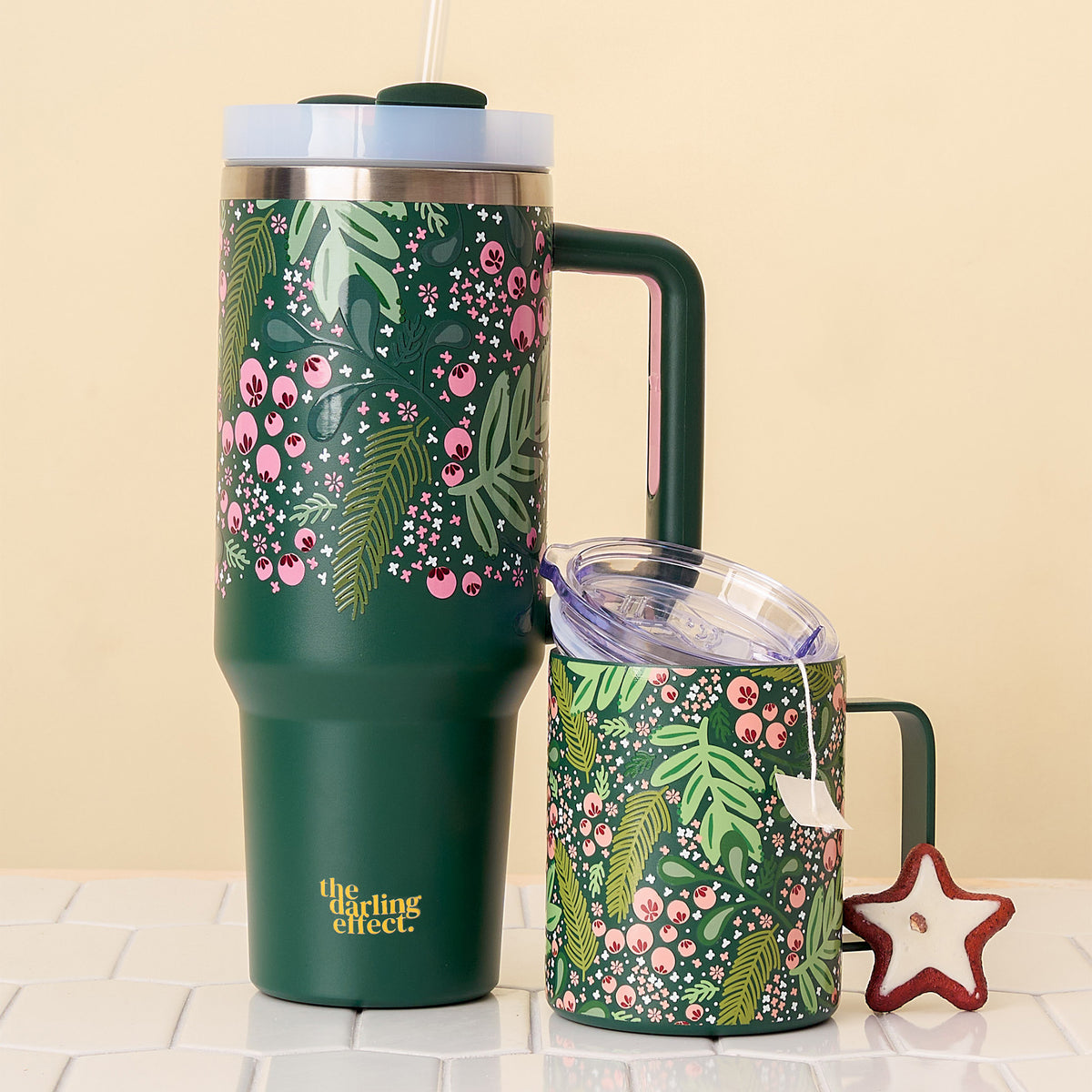 Two Jolly Sprig Green 40 oz Take Me Everywhere Tumblers by The Darling Effect, featuring a pink and white floral pattern, sit on a tiled surface beside a star-shaped cookie—perfect for holiday celebrations.