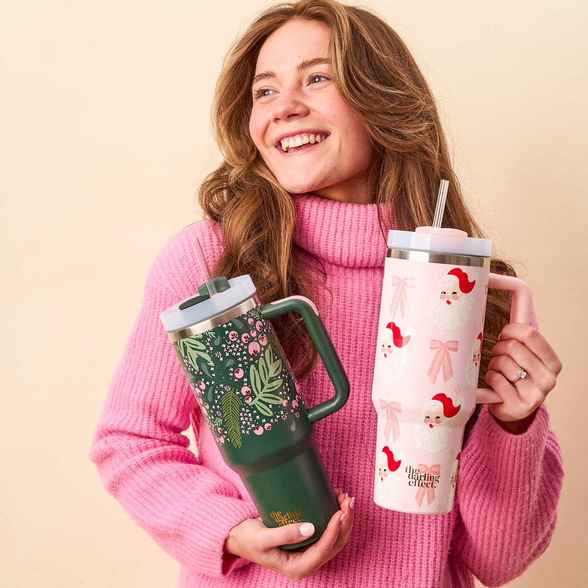 A smiling woman in a pink turtleneck holds two 40 oz The Darling Effect tumblers: the Jolly Sprig Green Take Me Everywhere Tumbler with a botanical pattern and a pink one with Santa faces and bows, perfect for the holidays.
