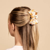 A woman with blonde hair, shown from the back against a beige background, styles her hair with The Darling Effect's Bouquet Beauty Sage Mini Satin Bow Claw Clip. She wears small hoop earrings and a white top.