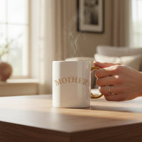 A hand holds The Darling Effect’s Mother 11 oz Cozy Cute Ceramic Mug, featuring “MOTHER” in gold letters and a golden handle. The steaming mug, ideal for coffee or tea, sits on a wooden table in a bright, inviting room.