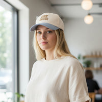 A smiling woman with long blonde hair stands against a plain beige background, wearing a white ribbed sleeveless top and The Darling Effect’s Boy Mom Classic Canvas Hat in white and blue. She holds the brim of her hat with one hand, effortlessly showcasing her casual style.