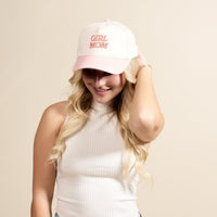 A smiling woman with long blonde hair wears a relaxed white top and lightly touches The Darling Effect’s Girl Mom Classic Canvas Hat, a light pink and white cap featuring “GIRL MOM” in pink letters, highlighting her effortless style.
