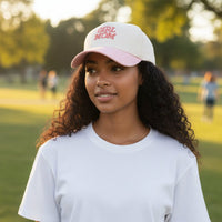 A woman with long curly hair wears a relaxed fit white t-shirt and The Darling Effect Girl Mom Classic Canvas Hat in white-and-pink. Outdoors in a park, she captures an effortless, casual style surrounded by trees and blurred figures.