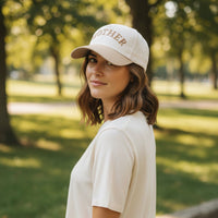 A woman with shoulder-length brown hair and a beige shirt wears The Darling Effect's Mother Classic Canvas Hat outdoors in a sunlit park, embracing an effortless casual style.