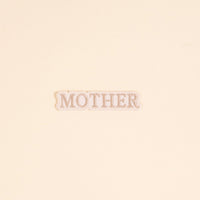 The Darling Effect’s Mother Mix & Match Iron-On Patch features “MOTHER” in tan serif on white with a scalloped edge. Perfect for customizing your items, it’s shown against a cream background.