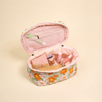 The-darling-effect's Bouquet Beauty Sage Glam & Go Quilted Cosmetic Bag, featuring a pink interior, is open on a beige background, displaying beauty essentials like a comb, brushes, lipstick, lip gloss, and a green tube.