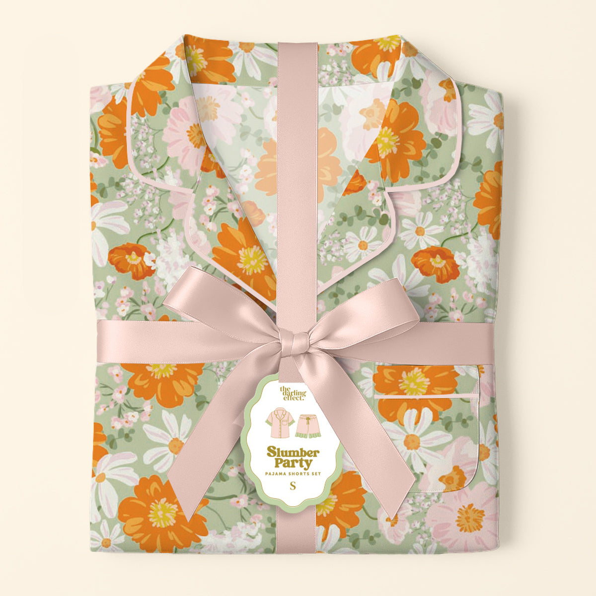 A folded, floral Bouquet Beauty Sage Slumber Party Pajama Shorts Set by The Darling Effect—crafted from ultra-soft fabric in green, orange, and white—is tied with a pale pink ribbon and labeled for a gift-ready surprise.