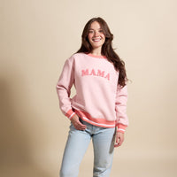 A woman with long brown hair smiles and poses with one hand in her pocket, wearing The Darling Effect's Mama Signature Crew in light pink with "MAMA" text and light blue jeans, standing against a beige background.