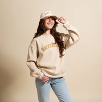 A smiling woman wears The Darling Effect’s Mother Signature Crew in beige with textured "MOTHER" letters, a matching cap, and light blue jeans. She stands against a beige background, holding her cap’s brim with one hand.
