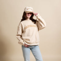 A young woman smiles in The Darling Effect’s Mother Signature Crew, a beige sweatshirt with “MOTHER” embroidery and a matching cap, crafted from soft fabric for everyday comfort.
