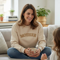 A smiling woman wears The Darling Effect's Mother Signature Crew beige sweatshirt with MOTHER on it, sitting cross-legged on a sofa, mug in hand. A small child is partially visible in the cozy, well-lit living room.