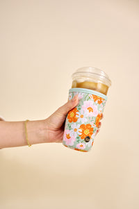 A hand holding an iced coffee in a clear cup with a plastic lid, wrapped in the-darling-effect Bouquet Beauty Sage Sip Savvy Drink Sleeve-Large, against a beige background.