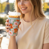 A woman in a cream-colored shirt smiles while holding an iced coffee outdoors on a sunny day, her cup wrapped in the-darling-effect’s Bouquet Beauty Sage Sip Savvy Drink Sleeve-Large, which features a stylish floral pattern.
