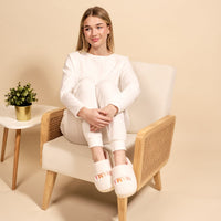 A woman in white loungewear hugs her knees and smiles, wearing The Darling Effect's Mama Fuzzy Slippers. She sits on a light chair beside a gold-potted plant on a white side table.